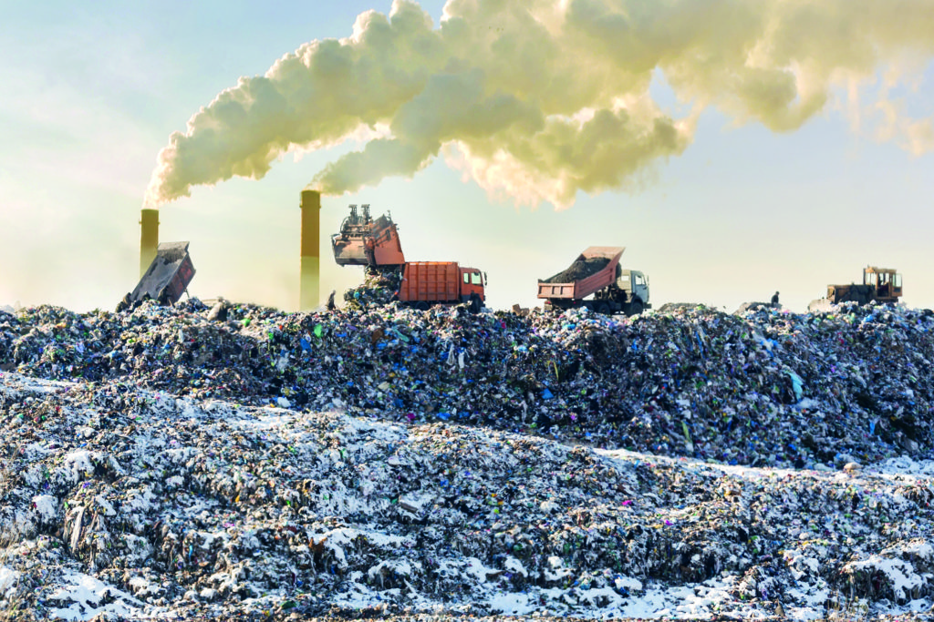 Dump trucks unloading garbage over vast landfill. Smoking industrial stacks on background. Environmental pollution. Outdated method of waste disposal. Survival of times past. Plastic & Climate: The Hidden Costs of a Plastic Planet.