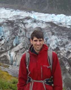 Image of a young man wearing a red jacket and backpack. Behind him is a glacier.