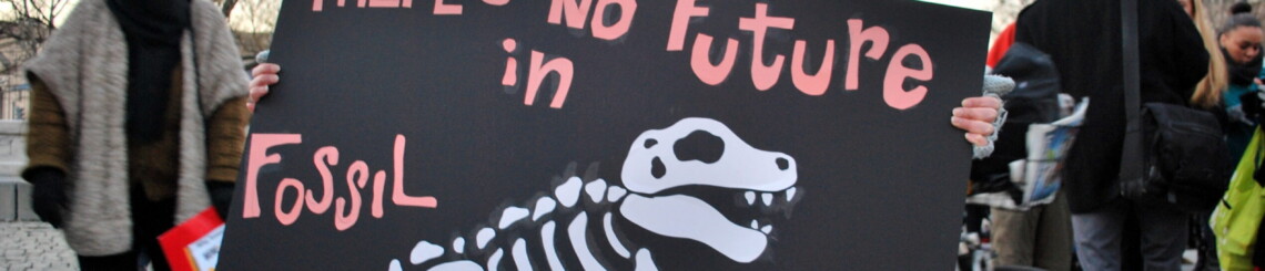 A young woman is at a protest. She carries a sign with "there's no future in fossil fuels" written in pink text and a t-rex skeleton.