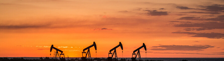 A silhouette of four oil wells are pumping oil. Behind the wells is a brilliant orange sunset.