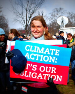 A woman smiles and stands at a protest holding a blue and red sign that says "Climate Action: It's Our Obligation"