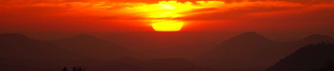 Brilliant orange sun setting into dark, tree-lined mountains. The sky is glowing red.